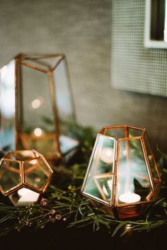 three glass lanterns sitting on top of a table next to greenery and candlesticks