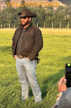 a man standing in the grass with his cell phone up to his chest and wearing a cowboy hat