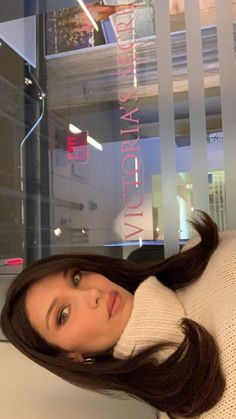 a woman with long brown hair wearing a white sweater and posing in front of a window