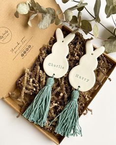 two ceramic rabbits with tassels are sitting in a box next to some leaves