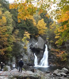 people standing on rocks near a waterfall in the woods with fall foliage around them and trees surrounding it