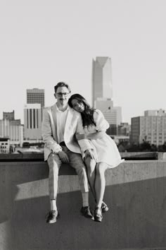 a man and woman sitting next to each other on top of a cement wall in front of a cityscape