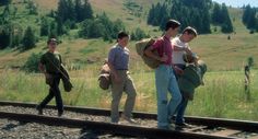 three boys are walking along the railroad tracks with backpacks on their backs and one boy is holding a book