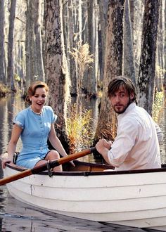 a man and woman in a row boat on the water with trees lining the shore