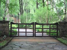 an iron gate in the middle of a stone walkway leading to a wooded area with trees