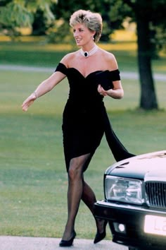 a woman in black dress walking next to a car