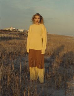 a woman standing in the middle of a field wearing a yellow sweater and brown pants