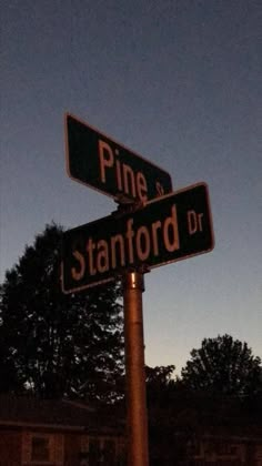 two street signs on top of a pole in front of some trees and buildings at night