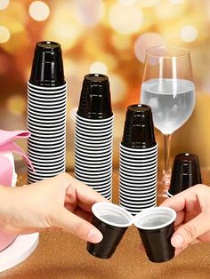 black and white paper cups are being held by two hands with wine glasses in the background