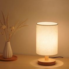 a table lamp sitting next to a white vase with flowers in it on a wooden stand