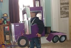 a young boy standing in front of a purple truck made out of cardboard and cardboard boxes