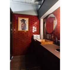 a bathroom with red tiles and a painting on the wall above the sink, in front of a round mirror