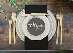 a place setting with black napkins and white plates, gold forks and silverware