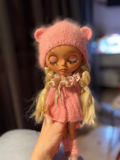 a doll with blonde hair wearing a pink dress and hat is being held up by someone's hand