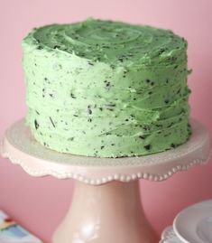 a cake with green frosting sitting on top of a white cake plate next to a pink wall