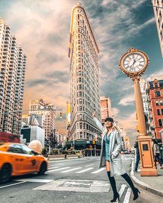 a woman crossing the street in front of tall buildings and a large clock on a pole