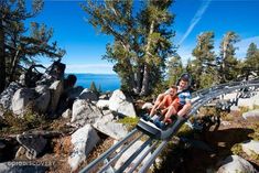 two people ride on a roller coaster over rocks and trees in the background, overlooking water