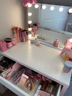 a white vanity with pink accessories and lights on the mirror above it is an open drawer