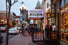 people are walking down the sidewalk in front of shops on a city street at dusk