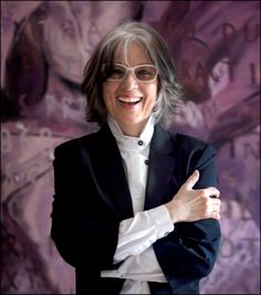 an older woman wearing glasses and a black jacket with her arms crossed, standing in front of a purple background