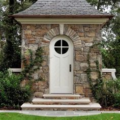 a small stone building with a white door and window in the center surrounded by greenery