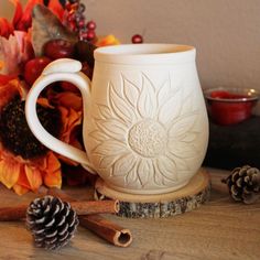 a white coffee mug sitting on top of a wooden table next to autumn leaves and cones