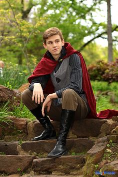 a young man dressed in medieval clothing sitting on some rocks and looking at the camera