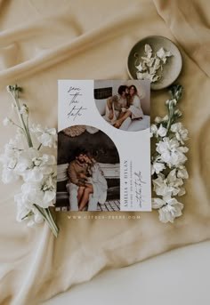 an image of a couple sitting on top of a bed next to flowers and a bowl