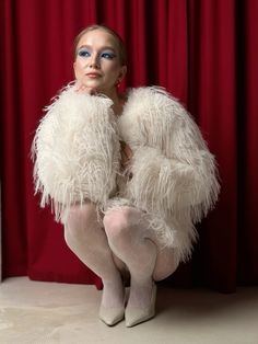 a woman with blue eyes and white feathers on her body sitting in front of a red curtain