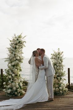 a bride and groom kissing in front of an archway with flowers on the beach behind them