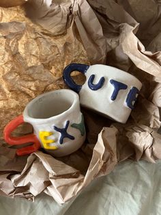 two ceramic mugs sitting on top of some crumpled up paper and one has the word put in it