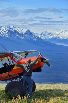 a man standing next to an airplane on top of a grass covered field with mountains in the background