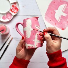 a woman is painting a cup with pink and white designs on the side, while she holds a paintbrush in her left hand