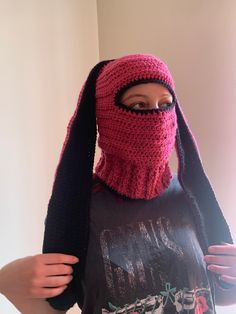 a woman wearing a pink knitted mask and black scarf over her head, standing in front of a white wall