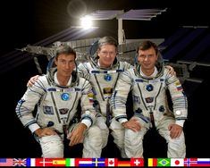 three men in space suits sitting next to each other with flags around them and an image of the international space station