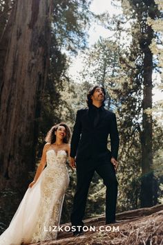 a bride and groom walking through the woods