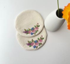 two coasters with embroidered flowers on them next to a white vase and orange flower