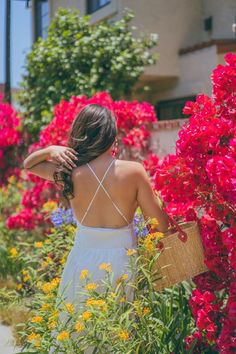 a woman in a white dress is walking through some flowers and holding a straw bag