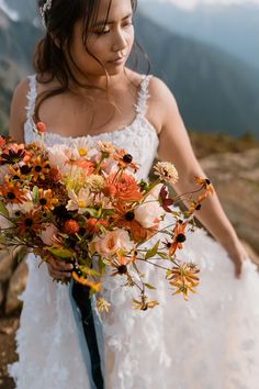 a woman in a wedding dress holding a bouquet of flowers on top of a mountain