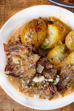 a white bowl filled with meat and potatoes on top of a wooden table next to a fork