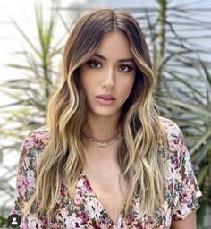 a woman with long blonde hair wearing a floral shirt and gold choker necklace, looking at the camera