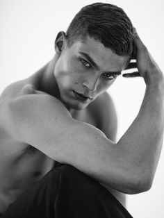 a shirtless young man sitting on the floor with his hands behind his head and looking at the camera