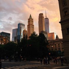 people crossing the street in front of some tall buildings at sunset or dawn with pink clouds