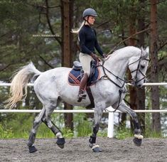 a woman riding on the back of a white horse in an outdoor arena with trees behind her