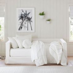 a white couch with pillows and blankets on it in front of two potted plants