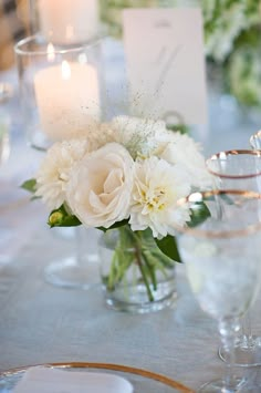 white flowers in glass vases sitting on a table with place cards and candles behind them