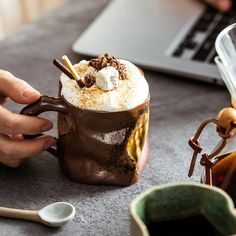 a person holding a coffee mug with whipped cream and chocolate toppings on it, next to a laptop