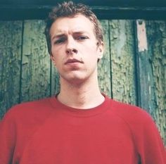 a man standing in front of a wooden wall wearing a red shirt and looking at the camera