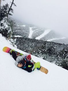 two snowboarders laying on the ground in the snow with trees and mountains behind them