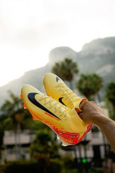 a person holds up a pair of yellow nike shoes in front of mountains and palm trees